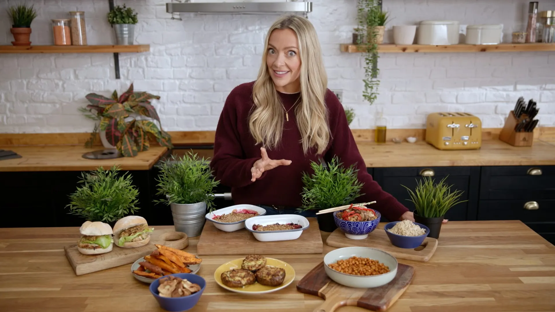 Long-haired blonde woman in a maroon jumper gestures behind a wooden table laid with sandwiches, bowls of grains, roasted vegetables and burgers, facing camera in a white-brick kitchen.