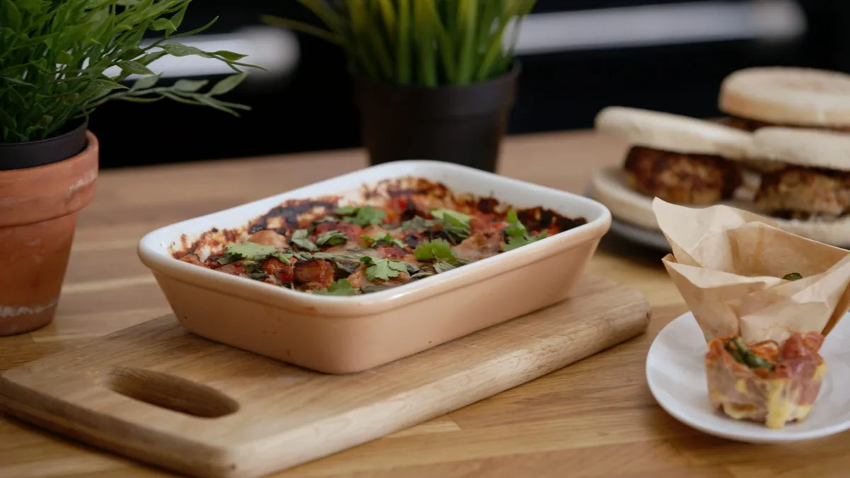 A dish of baked tomato-topped food with coriander in a rectangular baking dish on a wooden board, with potted plants, sandwiches and a pastry-wrapped canapé nearby.