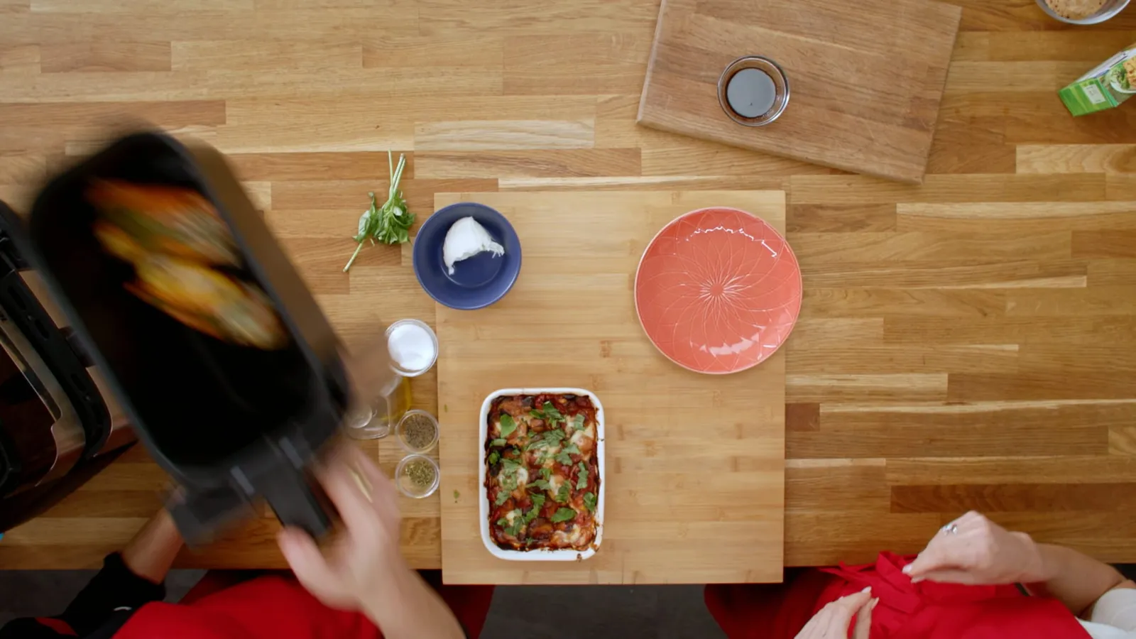 Overhead view of a wooden table with a baking dish of tomato and cheese bake, herbs, garlic and small bowls, as a person in red lifts a dark air fryer.