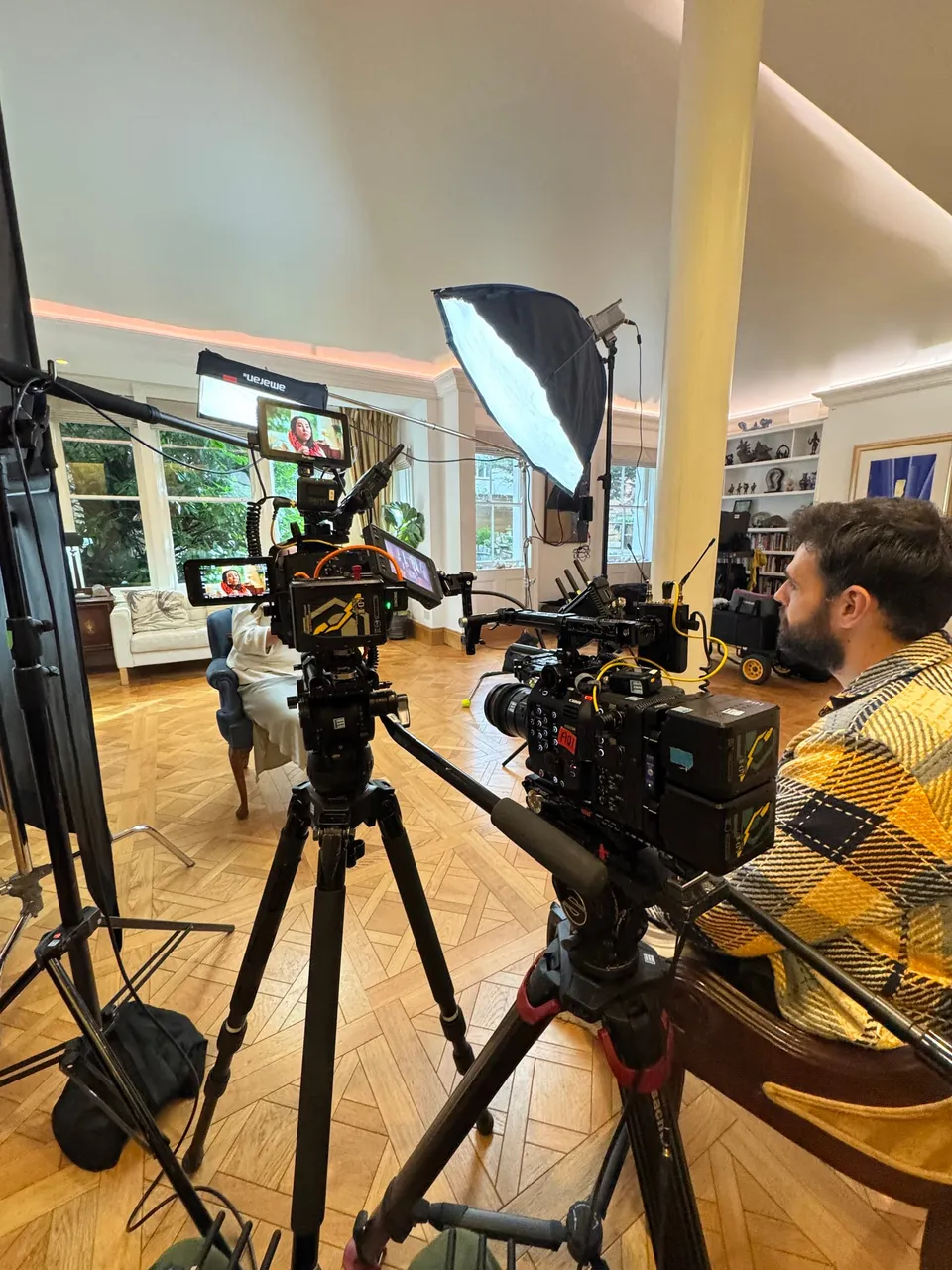 Two tripod-mounted cameras and a softbox light face a seated contributor by tall windows, a crew member watching a monitor in the foreground.