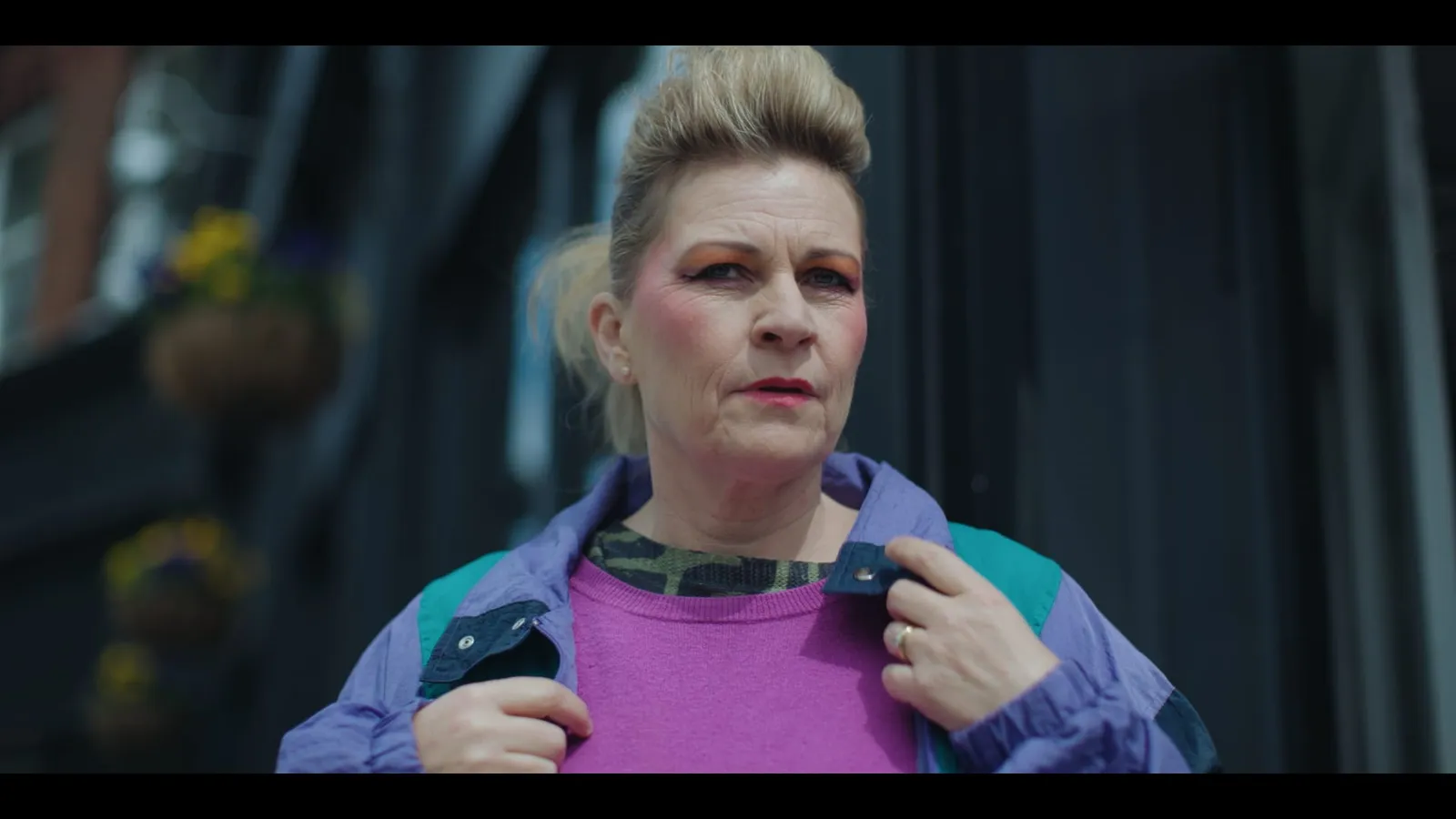 Close-up of a woman with blonde hair pulled up, bright eye makeup and a purple jacket, holding her collar and looking off-camera.
