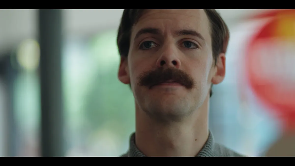 Close-up of a man with a moustache, facing camera with a neutral expression, set against a blurred background.