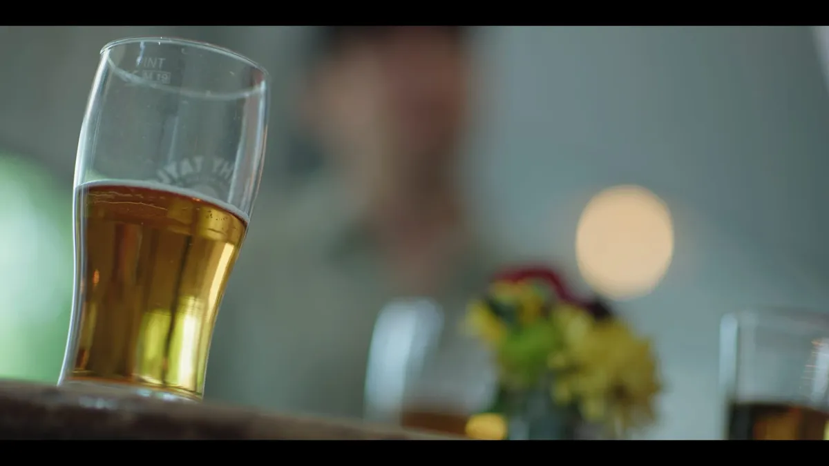 A part-filled pint glass in close focus on a pub table, with a blurred figure and flowers in the background.
