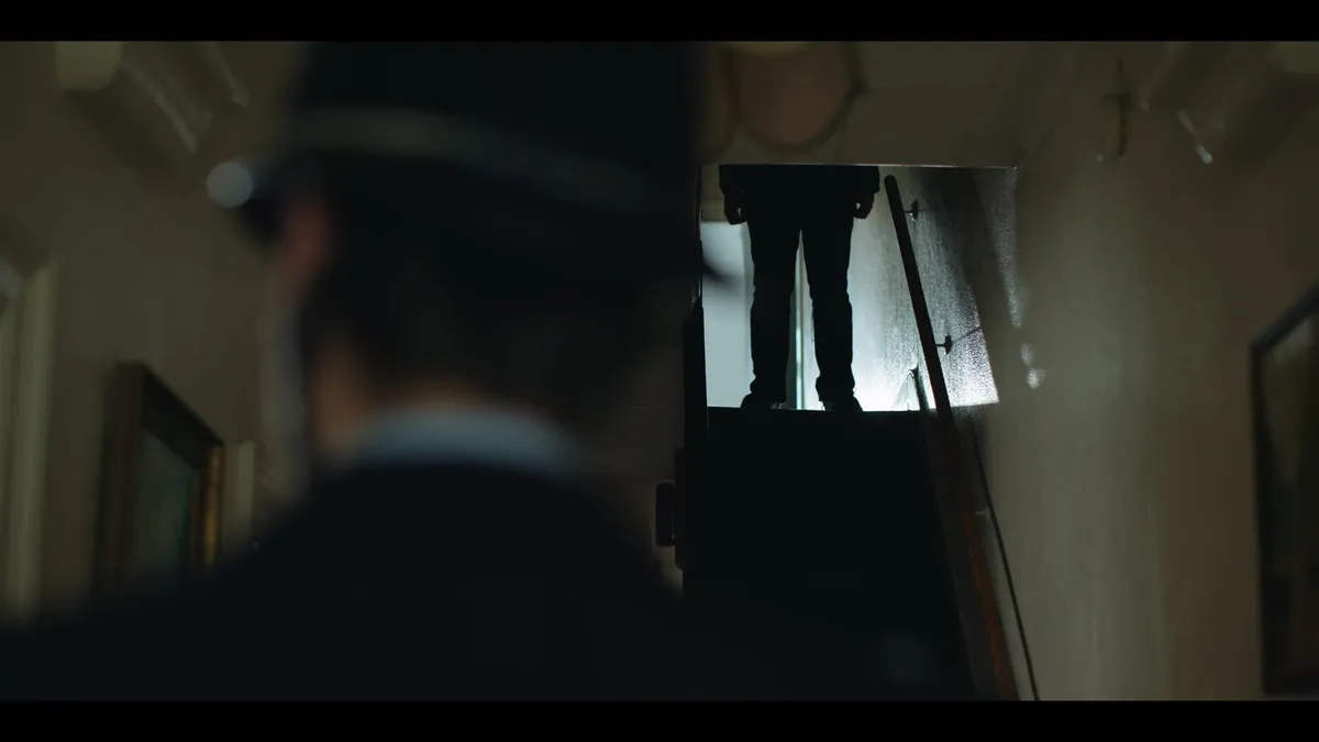 A blurred police officer in the foreground watches a figure's legs standing at the top of the stairs.