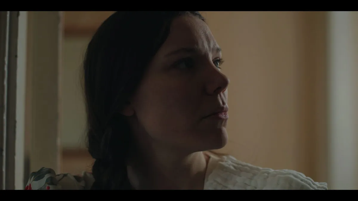 Close-up profile of a woman with dark hair, looking off-camera in a dimly lit room, soft light on her face.
