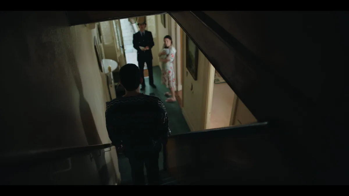 Seen from a stairway, a man faces down a hallway where a uniformed police officer and a woman in a floral robe stand near a front door, daylight entering behind them.