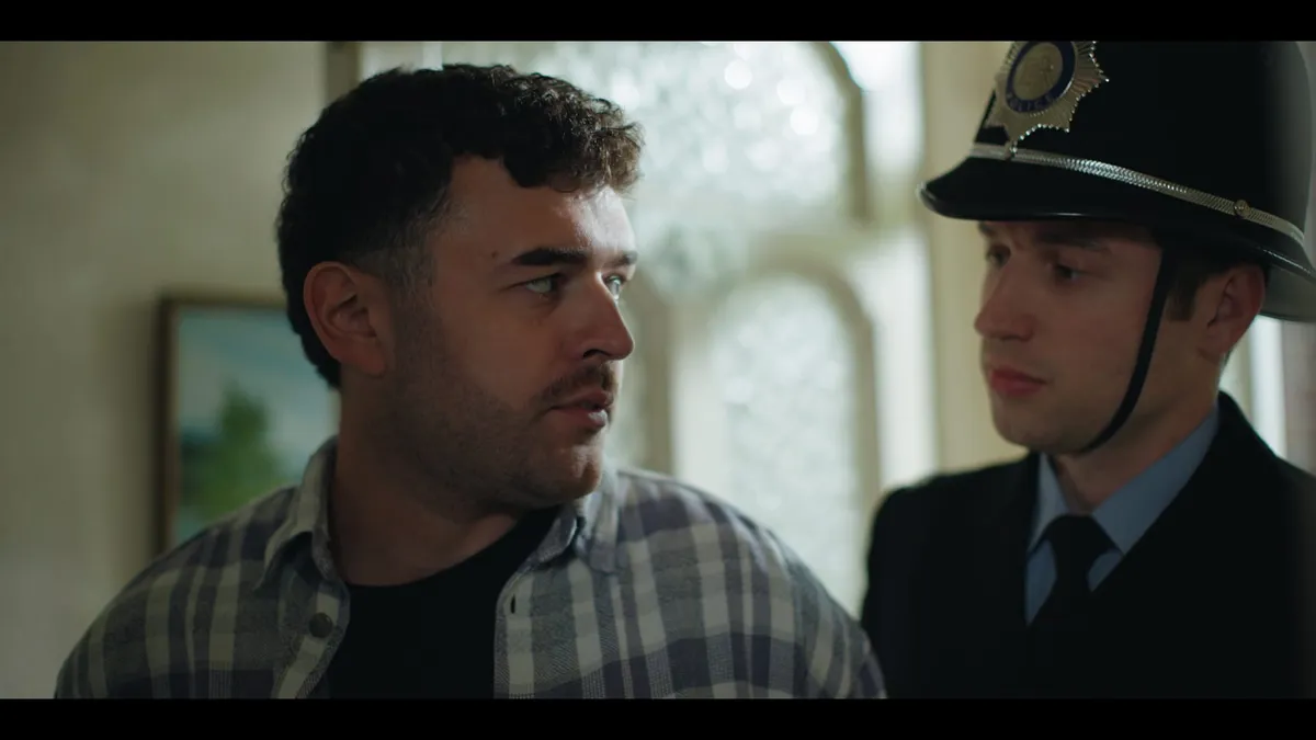 A man in a checked shirt looks in profile beside a police officer wearing a traditional custodian helmet, indoors near a leaded window.