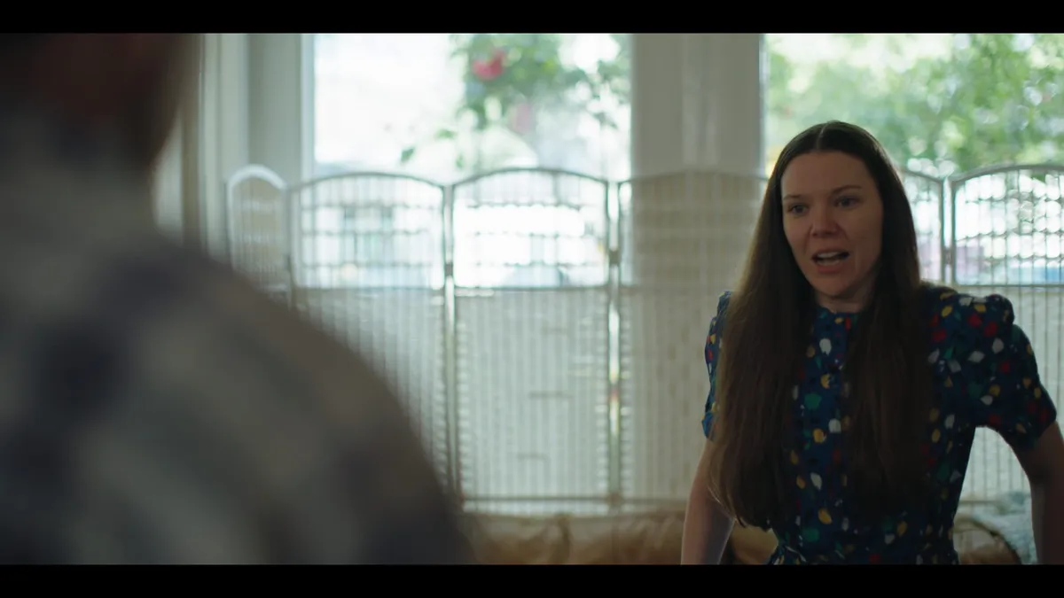 Woman in a floral dress speaking, facing an out-of-focus figure in the foreground, indoor room with patterned screen and windows behind her.