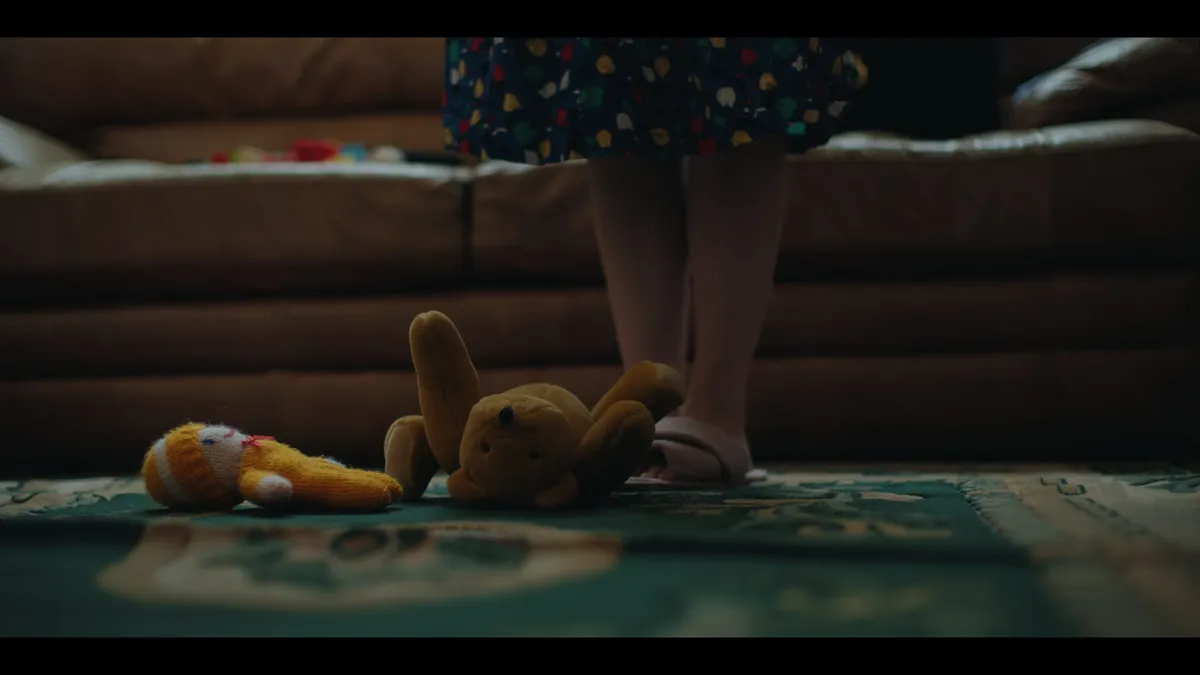 A woman's legs and sandals stand on a rug beside a stuffed toy and knitted doll, in front of a brown leather sofa.