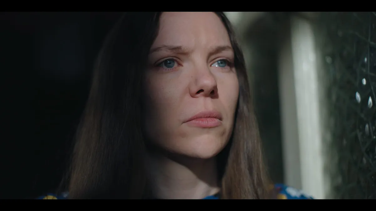 Close-up of a woman with long brown hair, looking off-camera towards a window, light falling across one side of her face.
