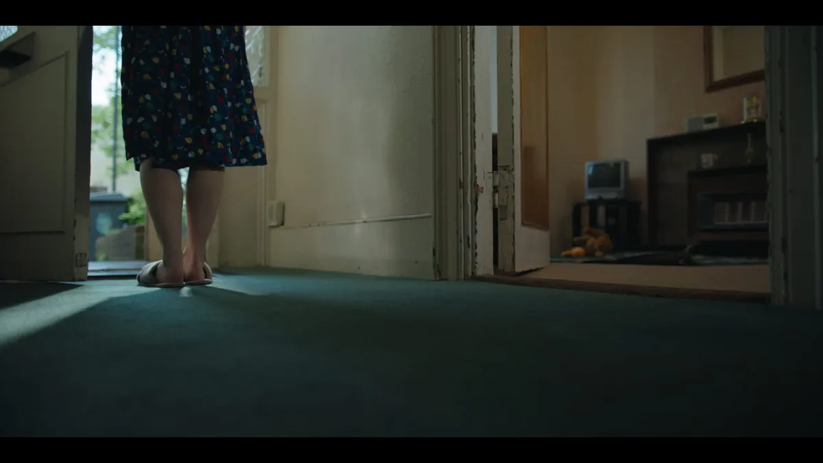 Low-angle shot of a woman's bare legs and sandals in a hallway, a patterned skirt visible, doorway opening to the front porch.