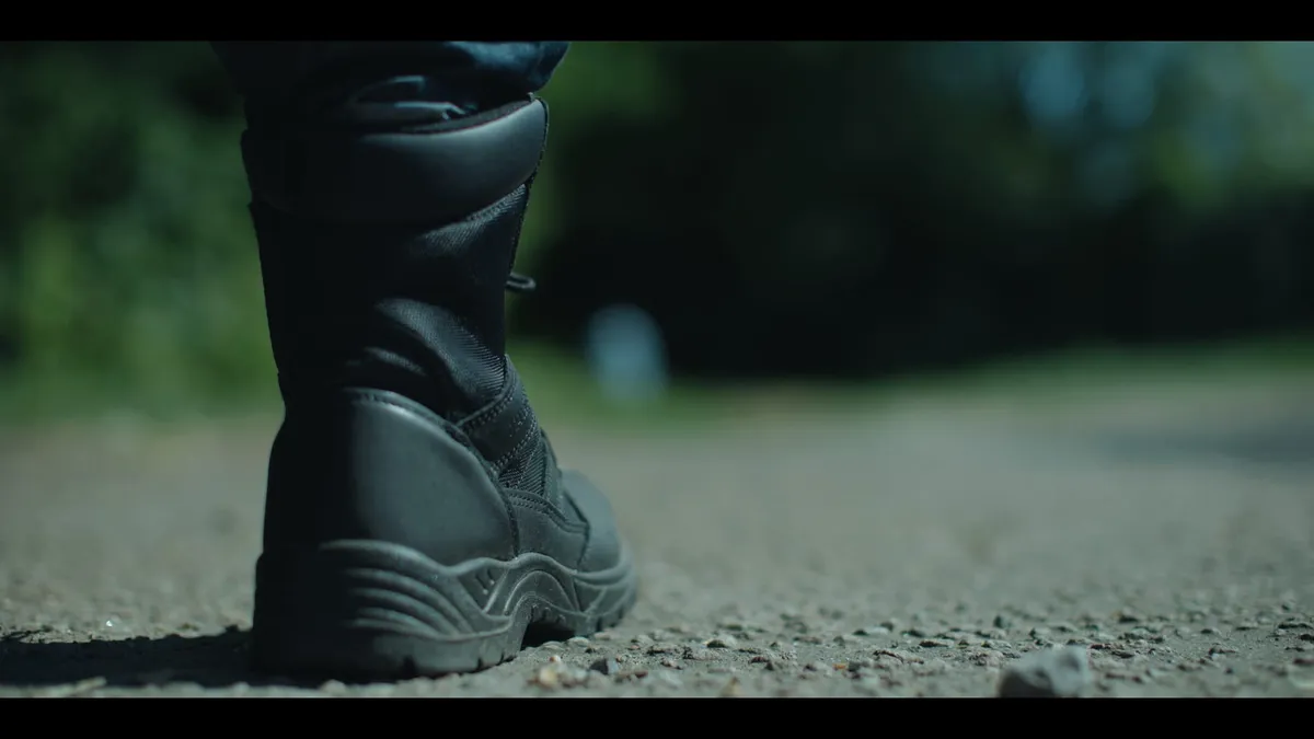Close-up of a black boot on gravel ground, low angle, with a blurred grassy path stretching into the background.
