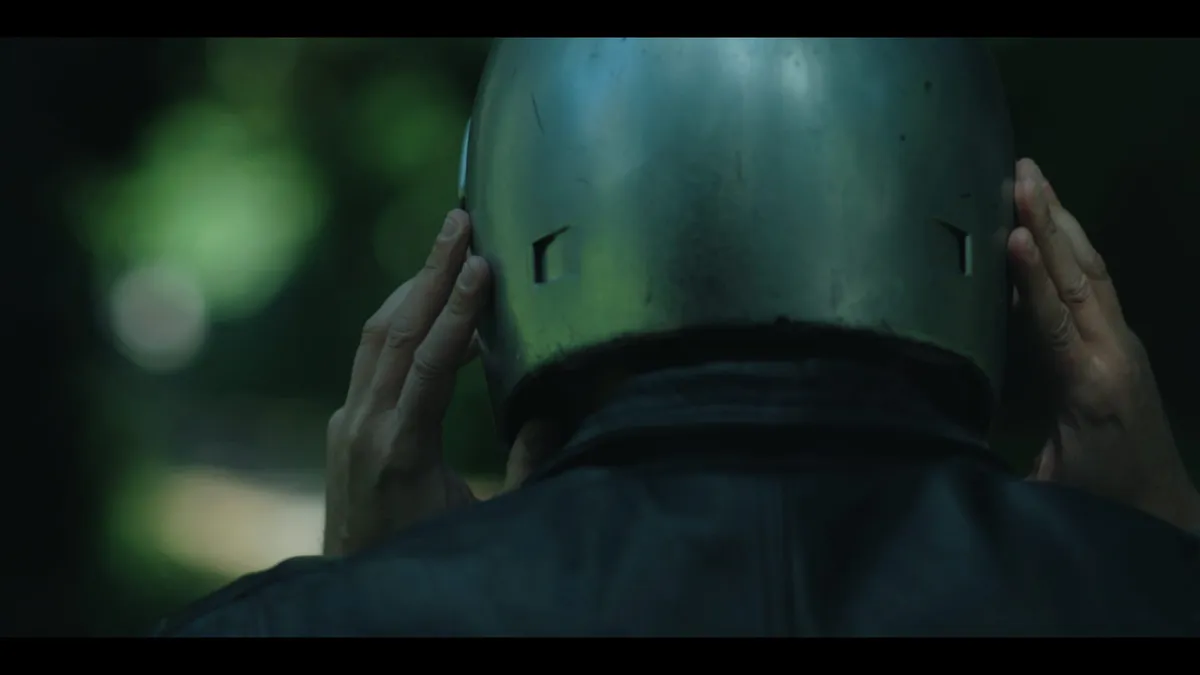 Close-up from behind of a person in a leather jacket adjusting a metal helmet with both hands, facing away from camera against blurred greenery.