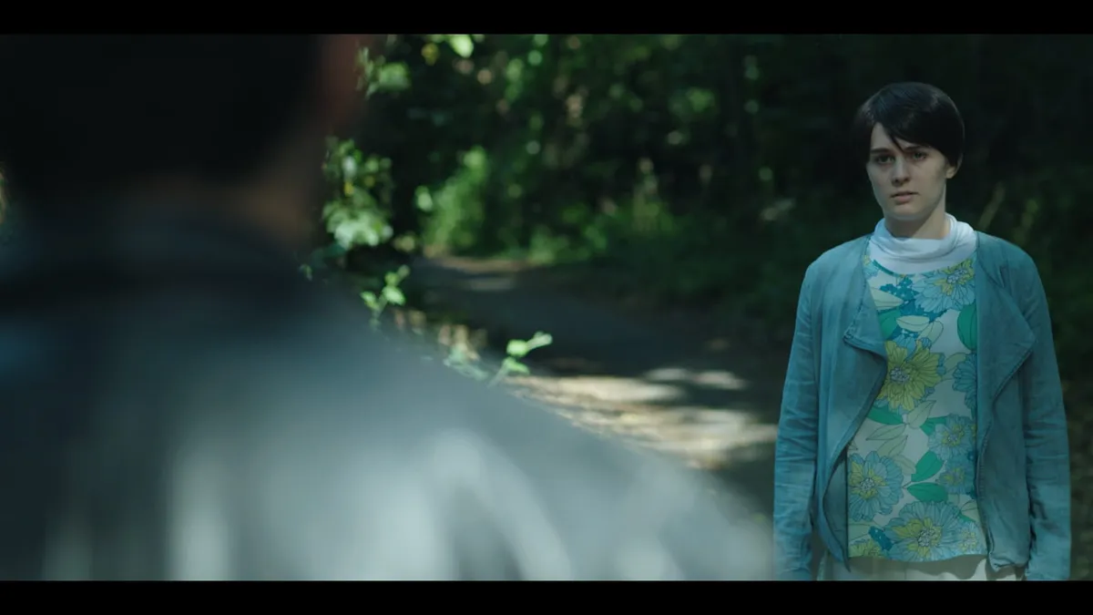 A young woman in a floral top and blue jacket stands on a wooded path, facing camera, an out-of-focus figure in the foreground.