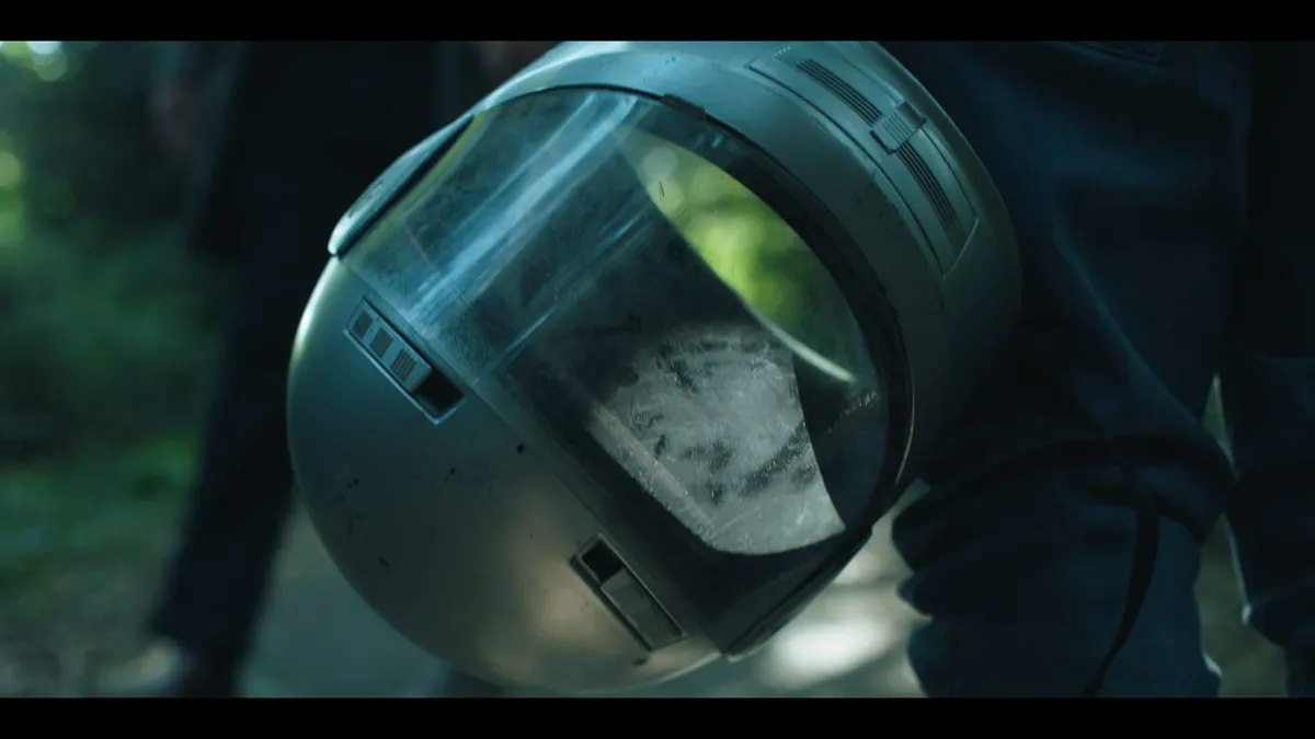 Close-up of a scuffed astronaut-style helmet held under one arm, visor smeared with dust, a figure blurred in woodland behind.