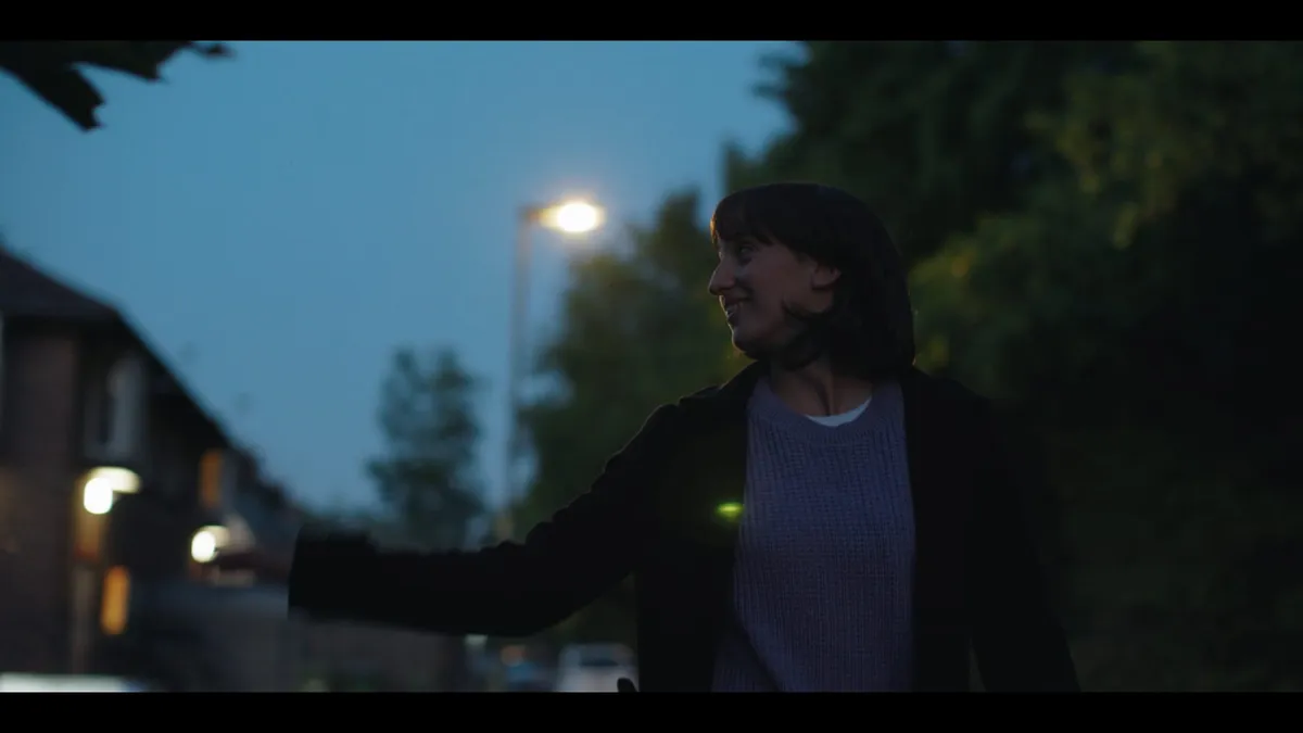 A woman in a dark coat walks a suburban street at dusk, smiling and looking off-camera, arm outstretched, a streetlamp lit behind her.