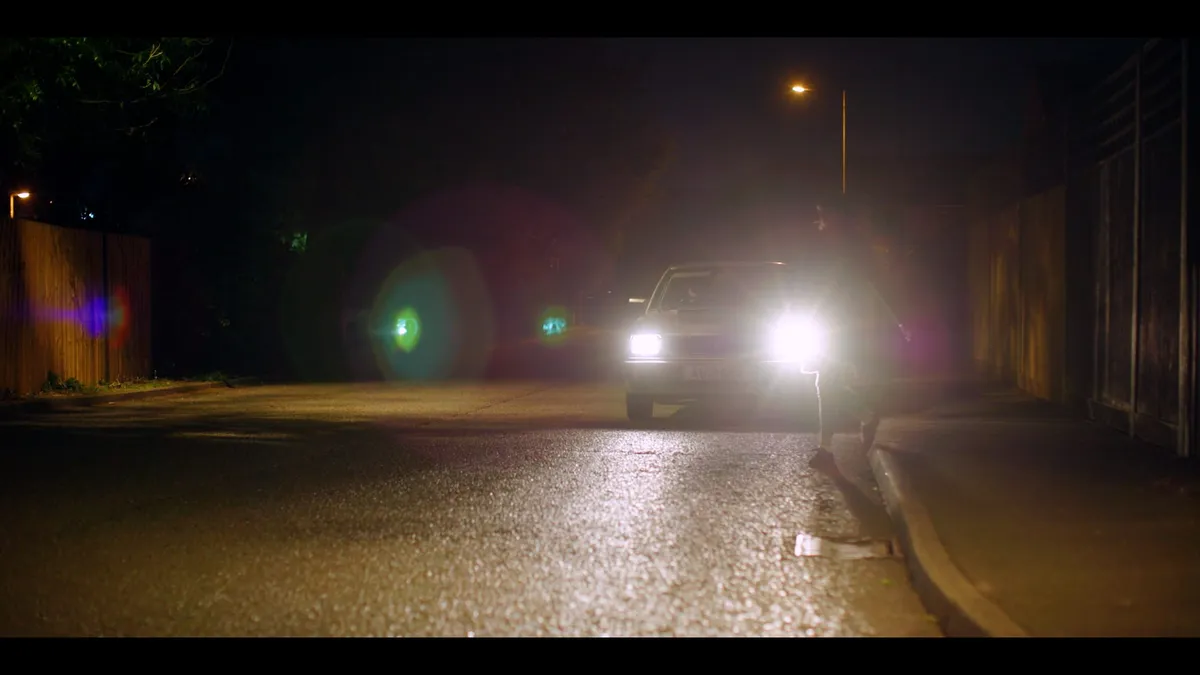 A car with headlights on faces camera on a dark, wet residential street at night, a figure standing beside it, streetlight glowing in the background.