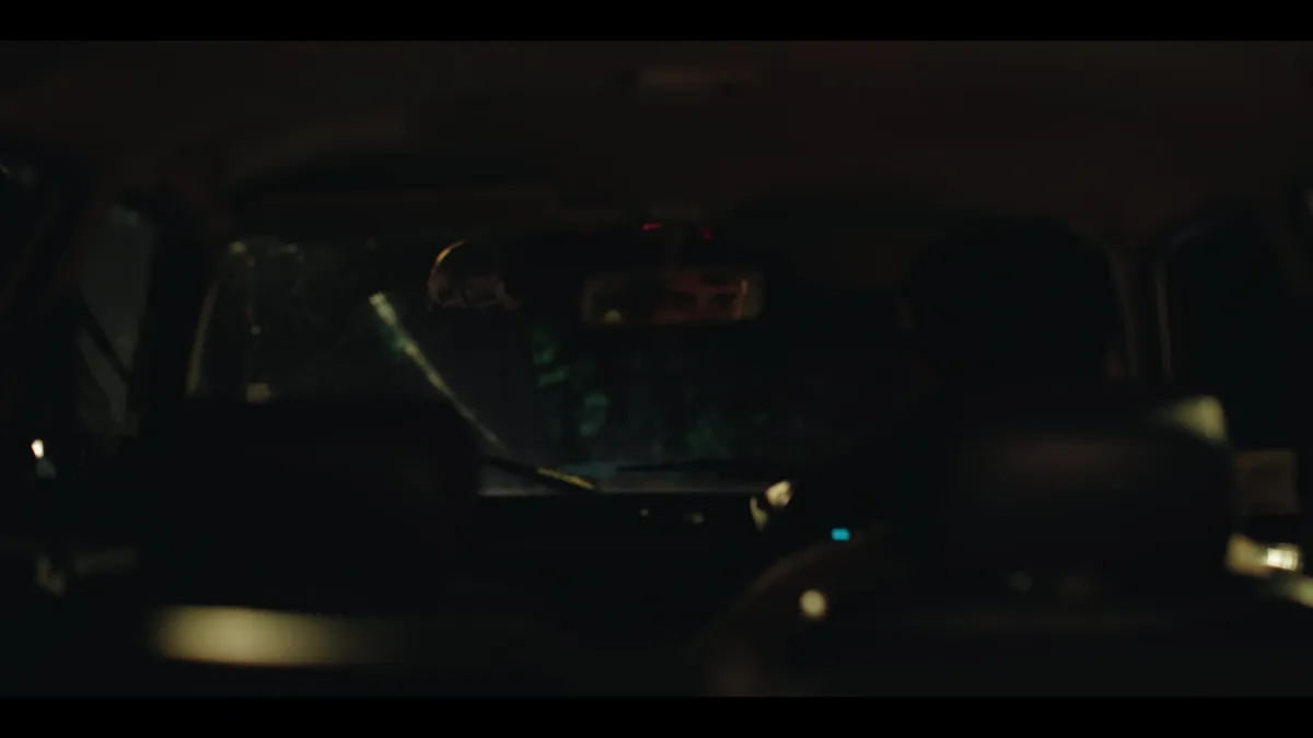 Interior of a car at night, seen from the back seats, a driver silhouetted ahead through the windscreen and rear-view mirror.