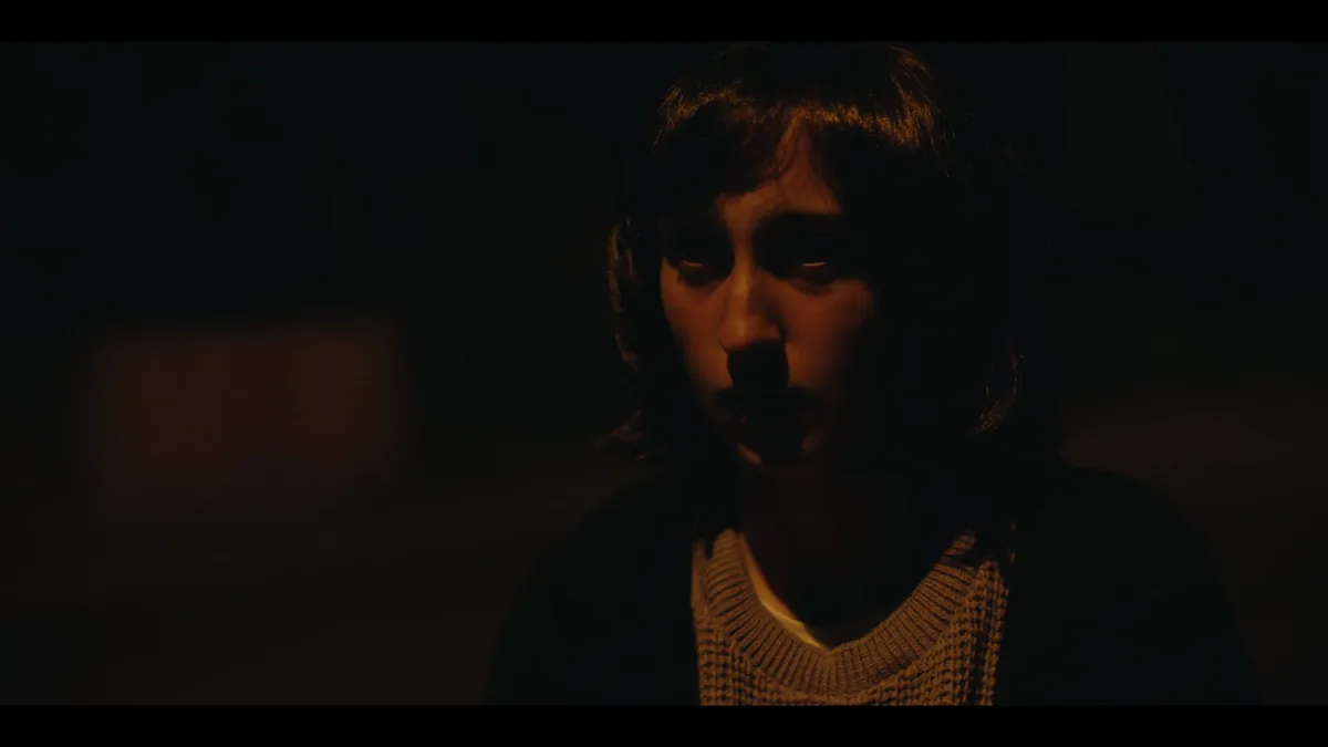 Close-up of a young woman with shoulder-length dark hair, dimly lit from below, facing camera in near darkness.