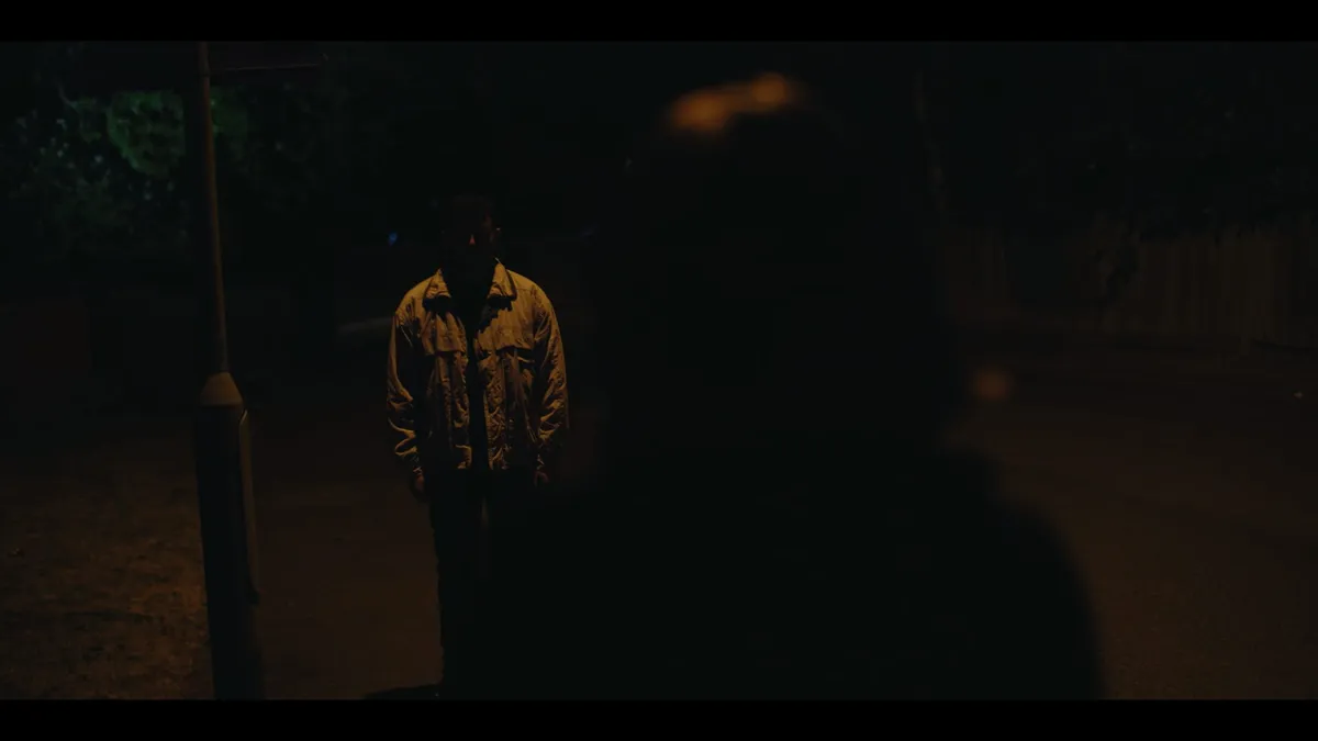 A man in a jacket stands facing camera on a dark street at night, lit by streetlight, an out-of-focus figure in the foreground.