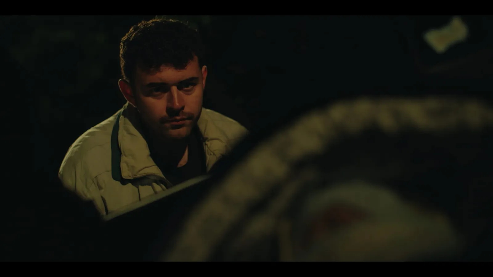 Night-time shot of a young man in a pale jacket looking off-camera, coiled rope blurred in the foreground, low side lighting against darkness.