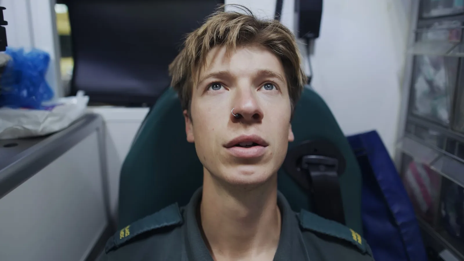A young man with short reddish-brown hair, in a dark green uniform, sits in an ambulance seat looking upward, close-up.