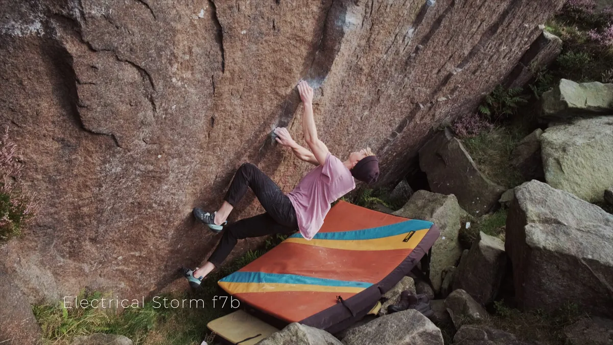 A climber in a pink t-shirt and dark cap grips a rock face overhang, crash pad below, boulders and heather surrounding.