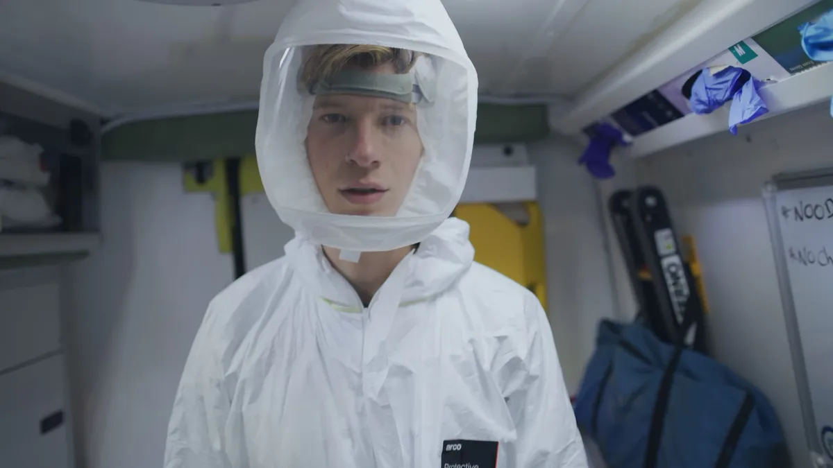 A young man with short reddish-brown hair wears a white protective suit with hood and visor, facing camera inside a vehicle interior.
