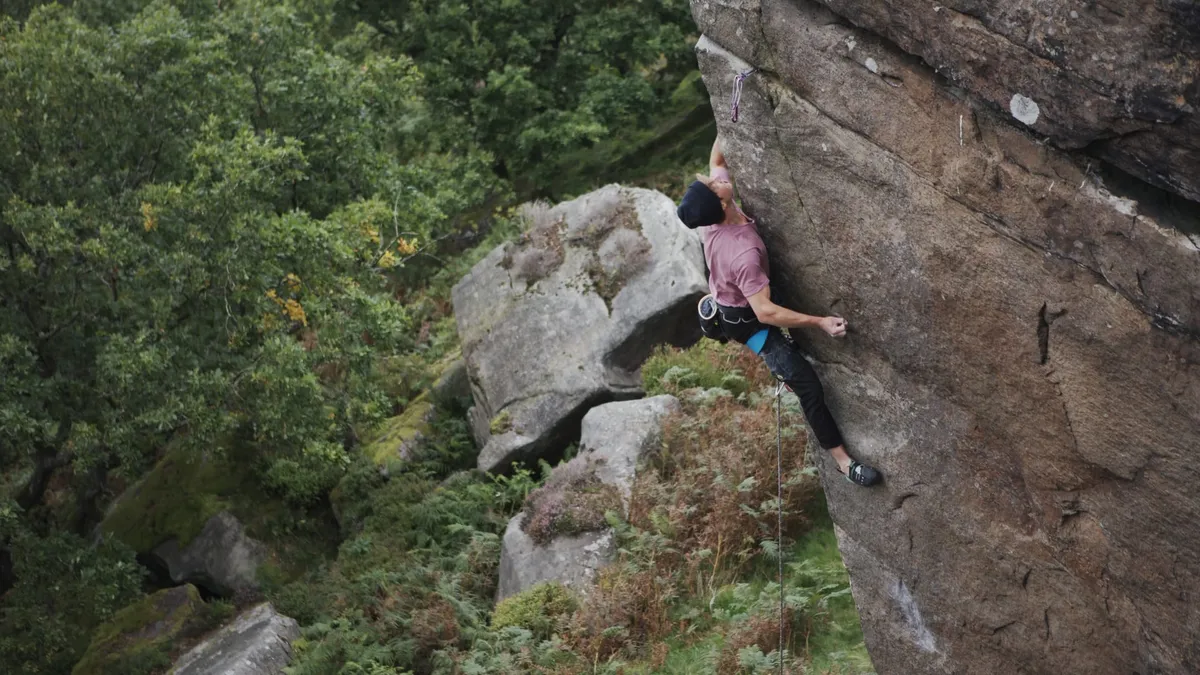 A climber in a pink t-shirt and dark cap scales a steep rock face, roped up, wooded hillside and boulders visible below.