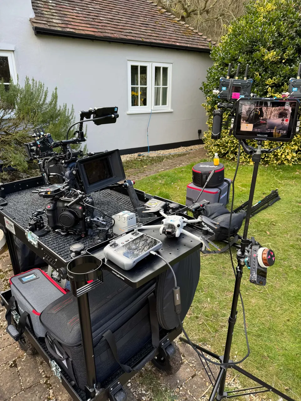 Camera trolley loaded with a cinema camera, monitors and a drone, set up on a garden lawn beside a white cottage.