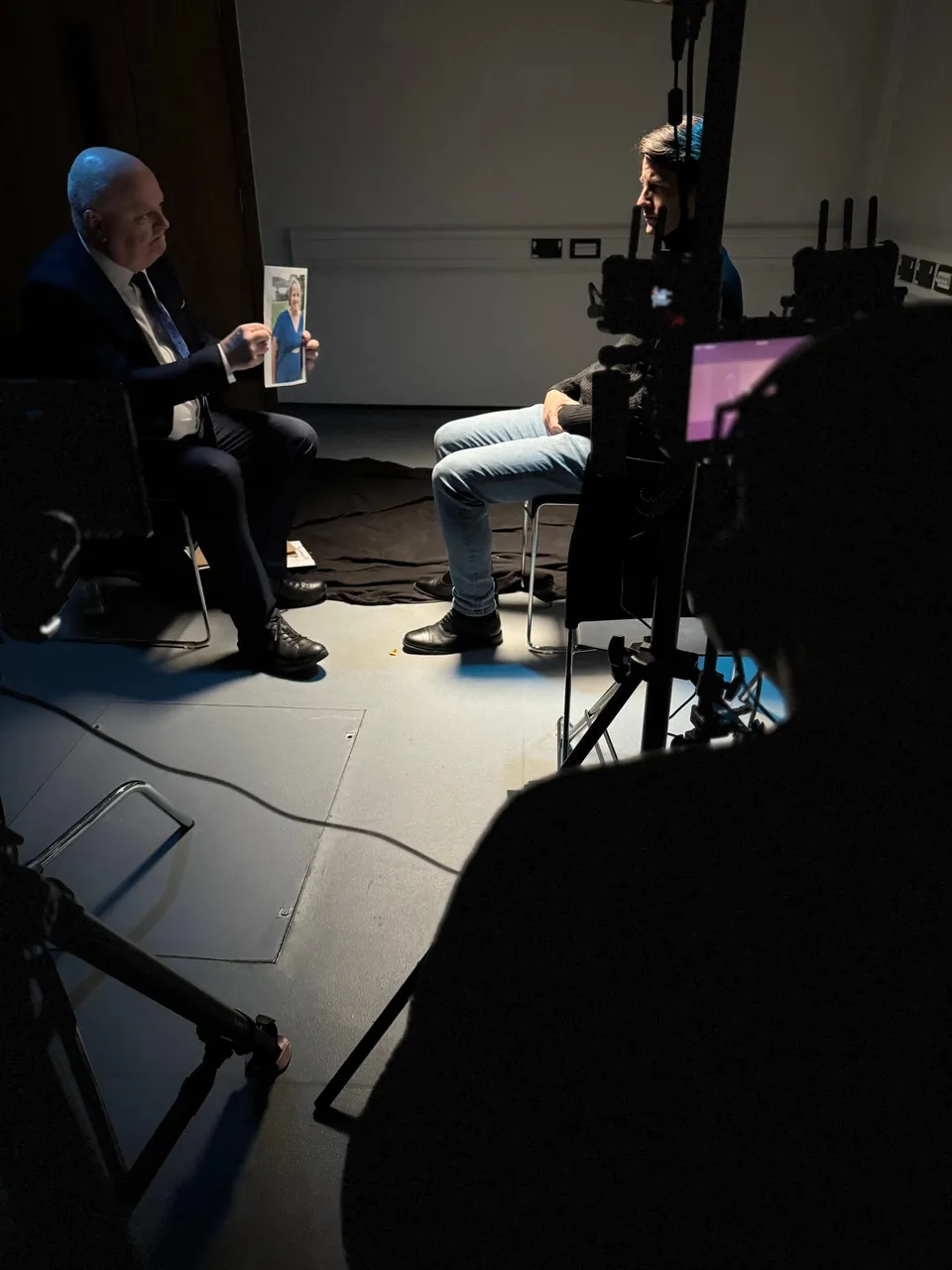 A seated man in a suit holds up a photograph while facing another man in jeans, filmed past camera and lighting rigs in a darkened studio.