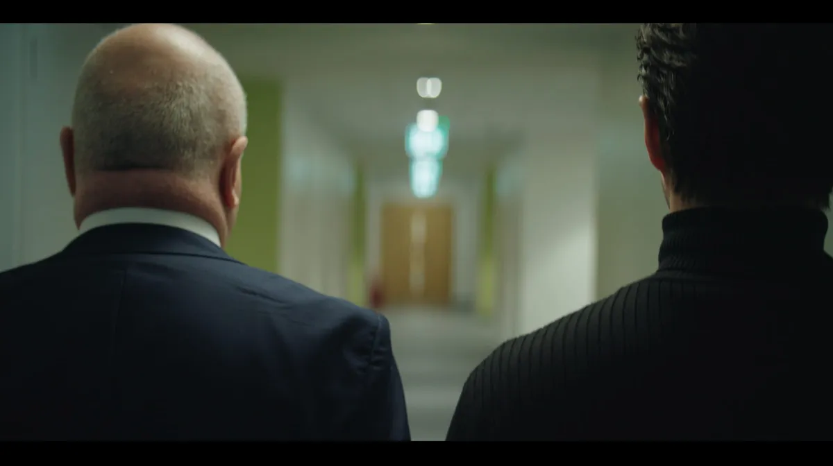 Two men seen from behind, walking down a corridor towards a lit exit sign and door, both out of focus in the background.
