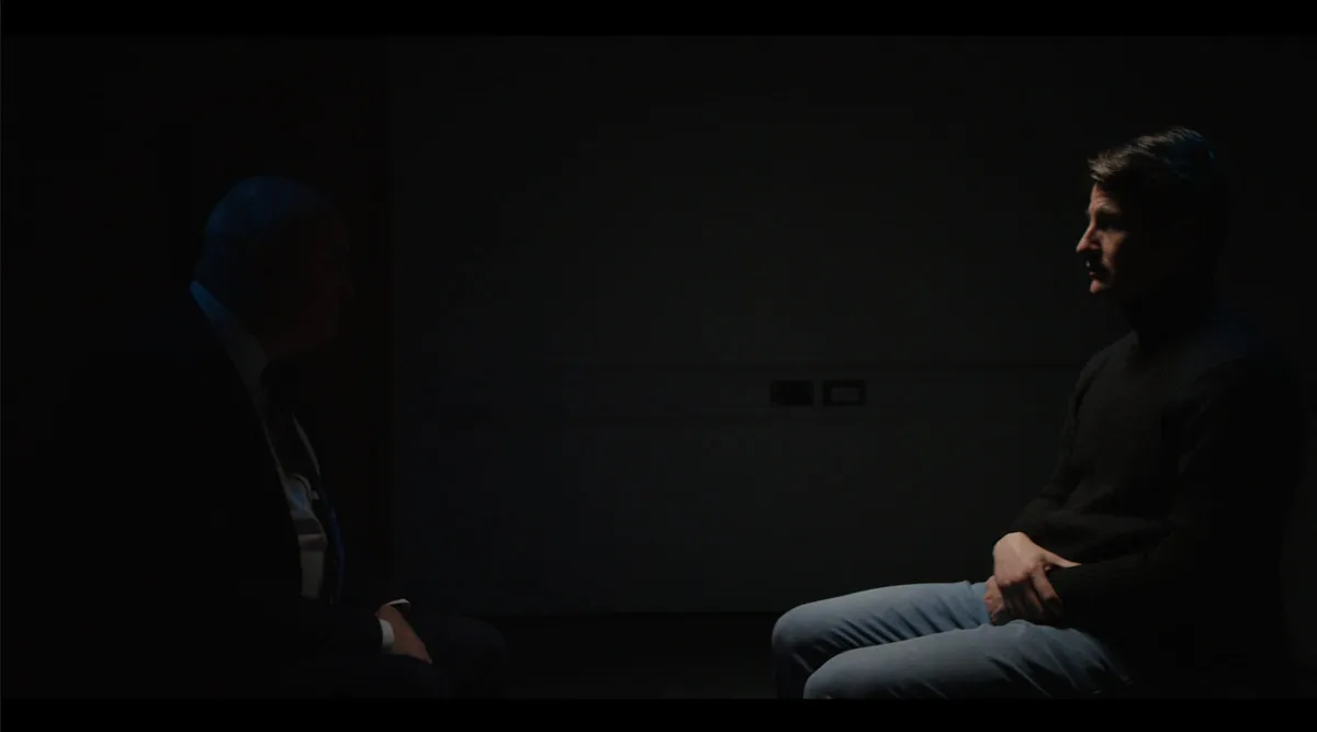 Two men seated in profile facing each other in a dimly lit room, an older man in a suit at left, a younger man in a jumper at right.