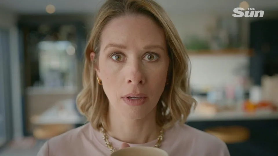 Close-up of a woman with wavy blonde hair, wearing a pink top and a gold chain necklace, facing camera in a kitchen.