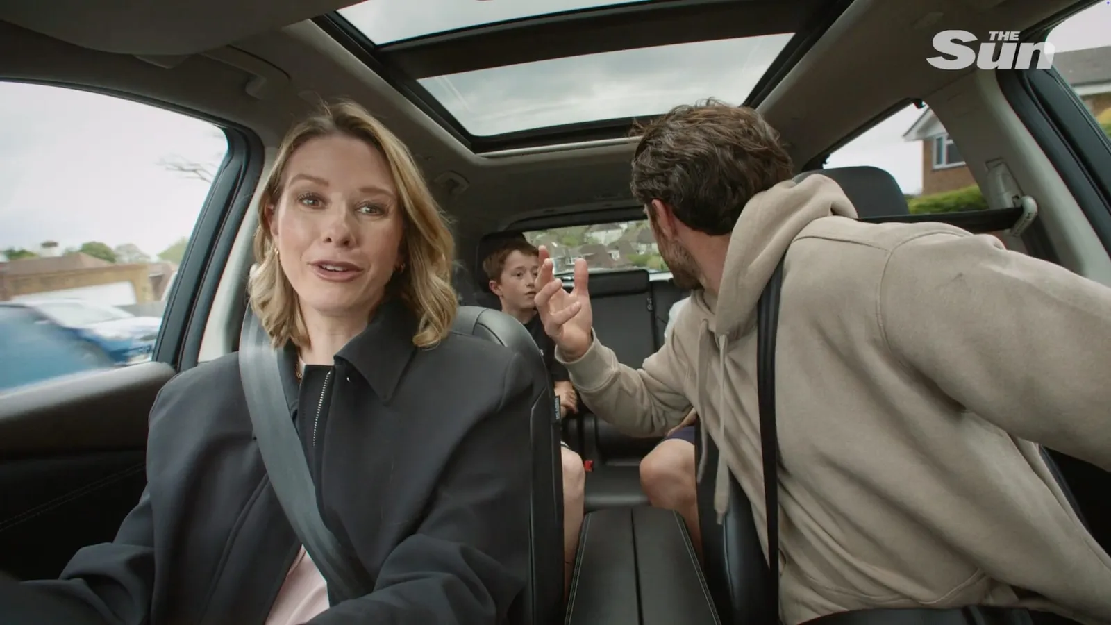 A woman with wavy blonde hair, in a black coat, sits in a car facing camera as a man in a beige hoodie gestures beside a boy in the back seat.