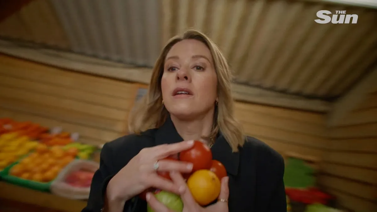 A woman with wavy blonde hair, in a black coat, holds tomatoes and a lemon at a market stall, facing camera.