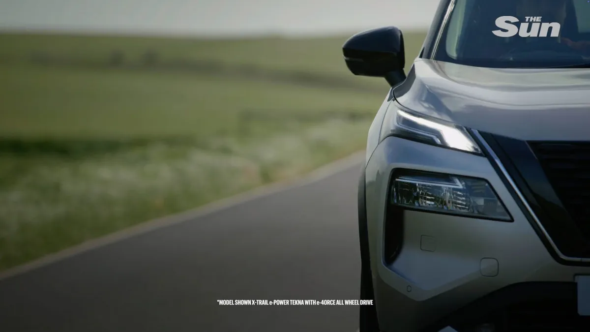 Close-up of the front wing, headlight and wing mirror of a silver car parked on a rural road beside green fields.