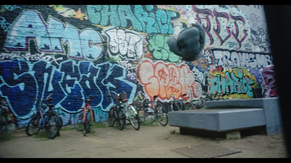 A person mid-somersault above concrete blocks, in front of a graffiti-covered wall with parked bicycles lined against it.
