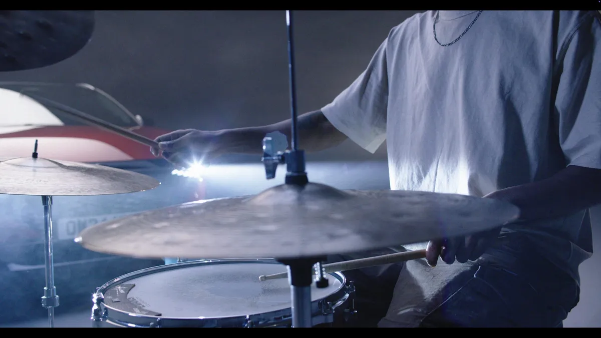 Close-up of a person in a pale grey T-shirt playing a drum kit, cymbals in foreground, a car and bright light visible behind in a smoky studio.