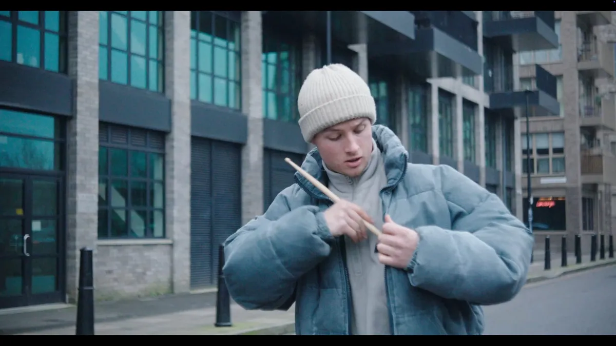 A young man in a cream beanie and blue puffer jacket holds a drumstick to his chest, standing on a street lined with modern buildings.