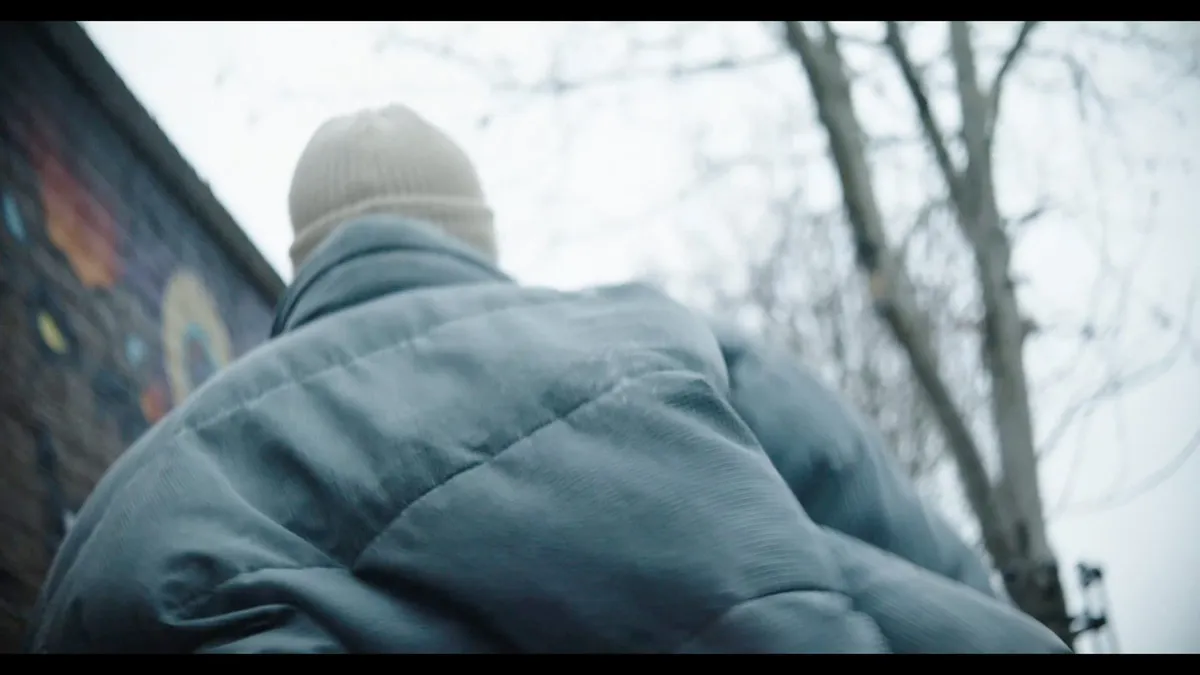 Low-angle view from behind of a person in a cream beanie and blue puffer jacket, walking beneath bare tree branches beside a graffiti-covered wall.