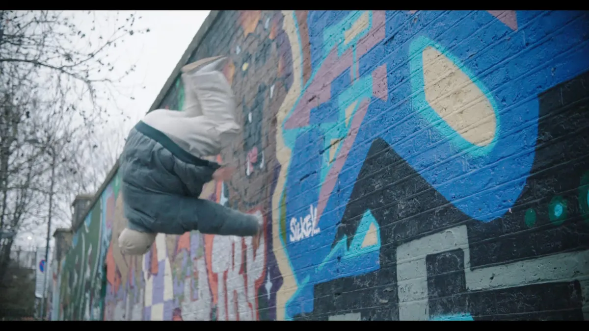 Low-angle view of a person in a cream beanie and grey jacket mid-somersault against a graffiti-covered wall, bare tree branches above.