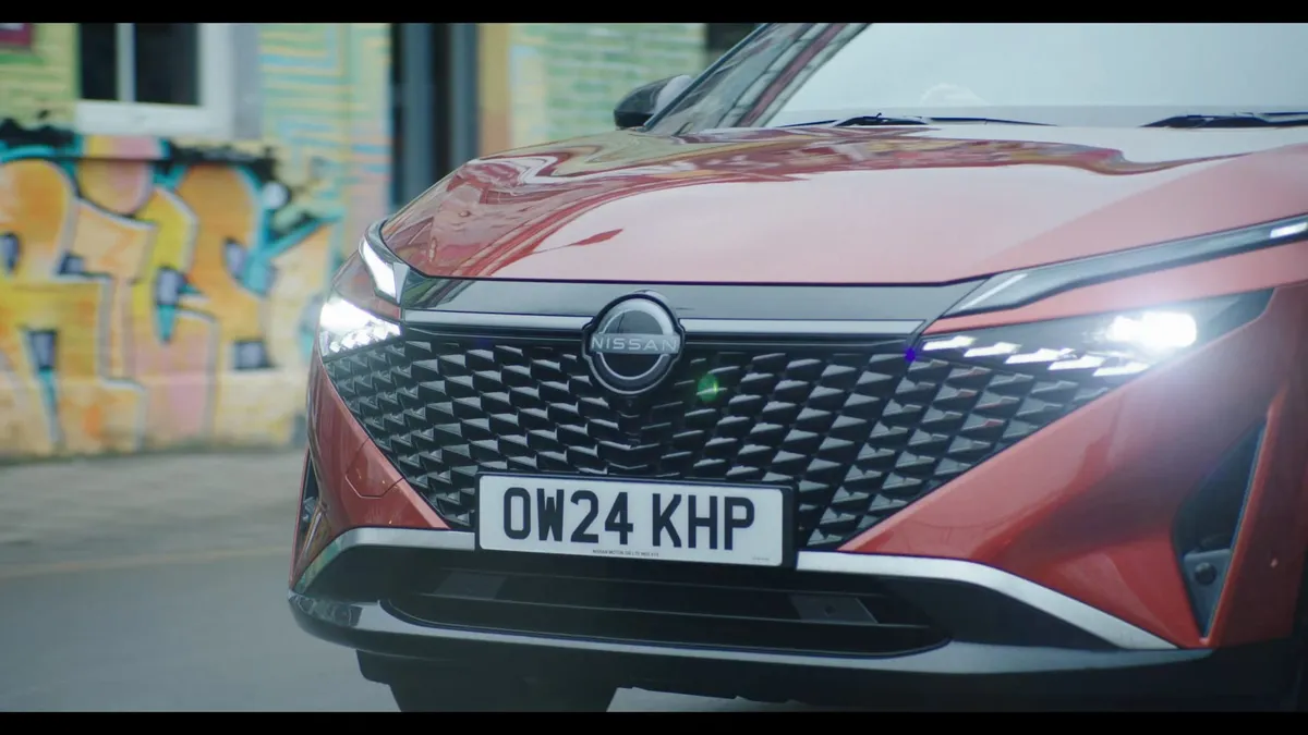 Close-up of a red car's headlight and grille with registration plate OW24 KHP, parked outside against a graffiti-covered wall.