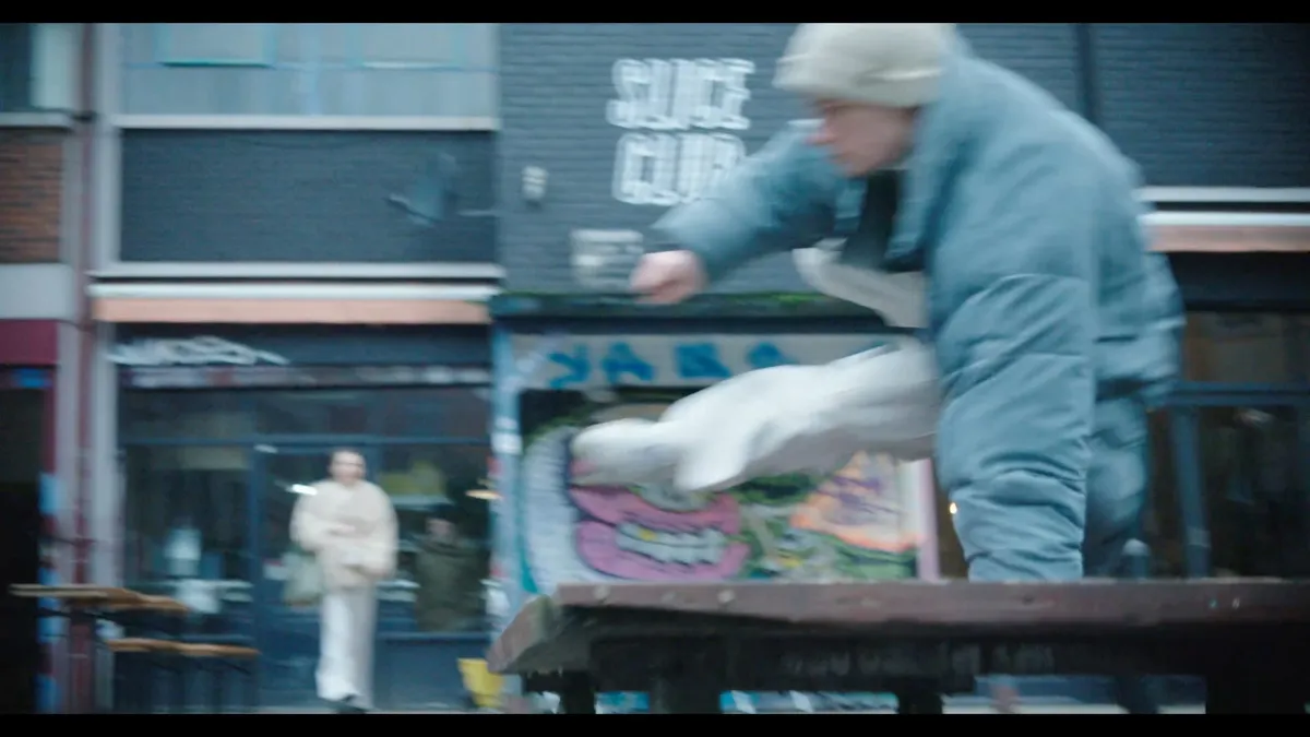 A person in a cream beanie and blue puffer jacket leaps off a raised platform, a shopfront and another figure blurred in the background.