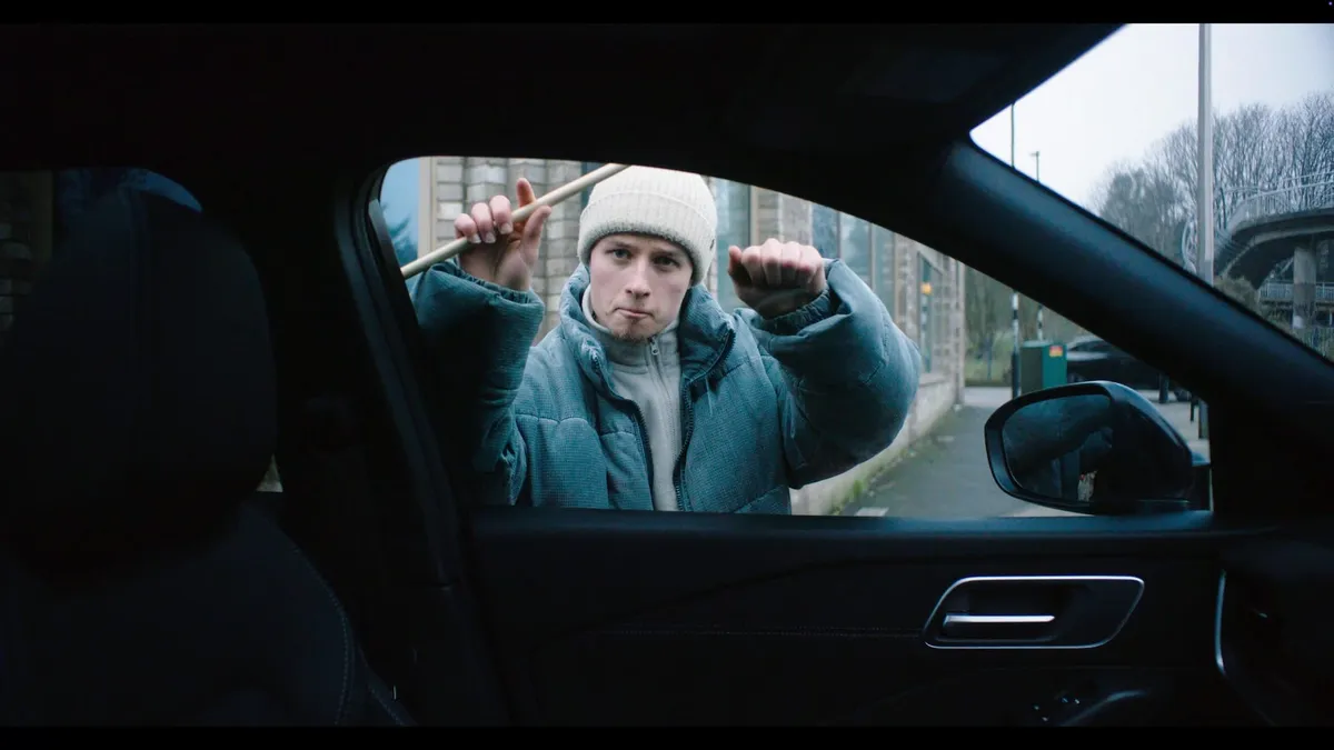 A young man in a cream beanie and blue puffer jacket raises fists and a drumstick, seen through a car's open window on a street.
