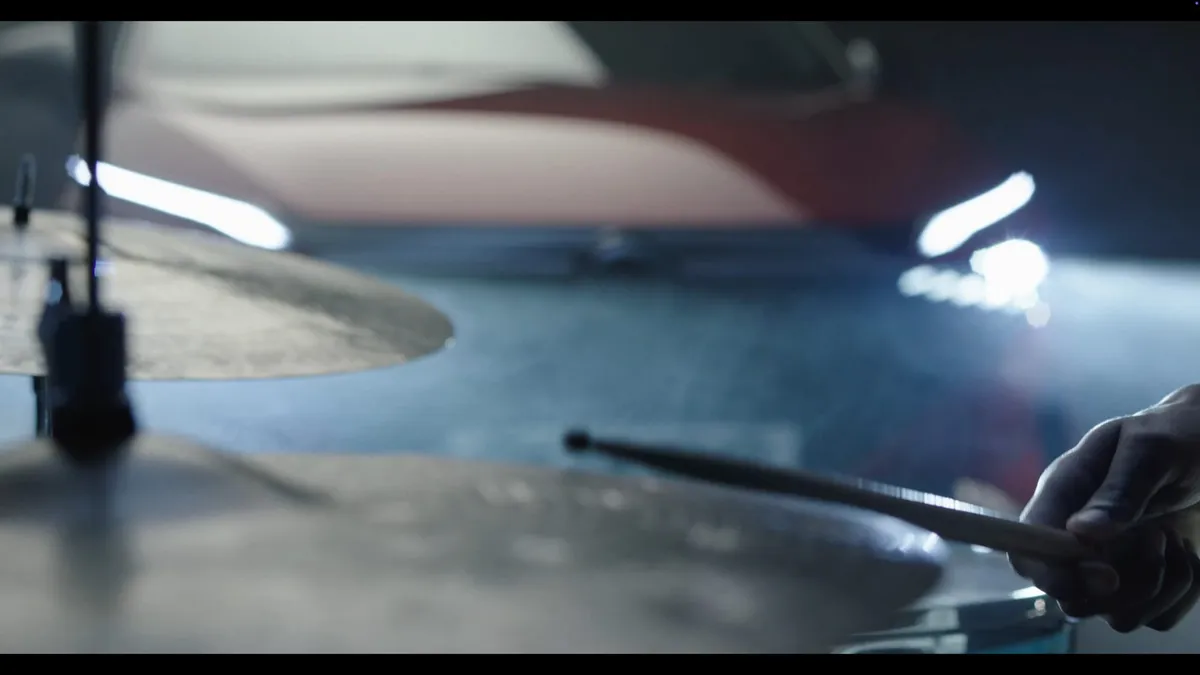 Close-up of a hand gripping a drumstick over cymbals, a red car and bright light visible behind in a smoky studio.