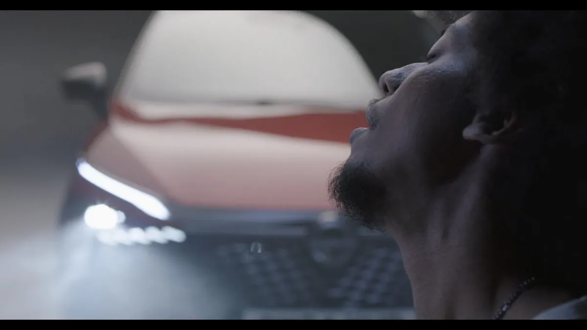 Close-up profile of a man with short curly hair and a beard, looking upward, a red car's raised bonnet and headlight glowing behind him in a smoky studio.