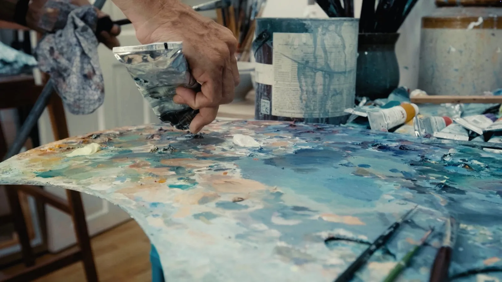Close-up of a hand squeezing paint from a tube onto a heavily loaded palette, brushes and paint tubes visible behind.
