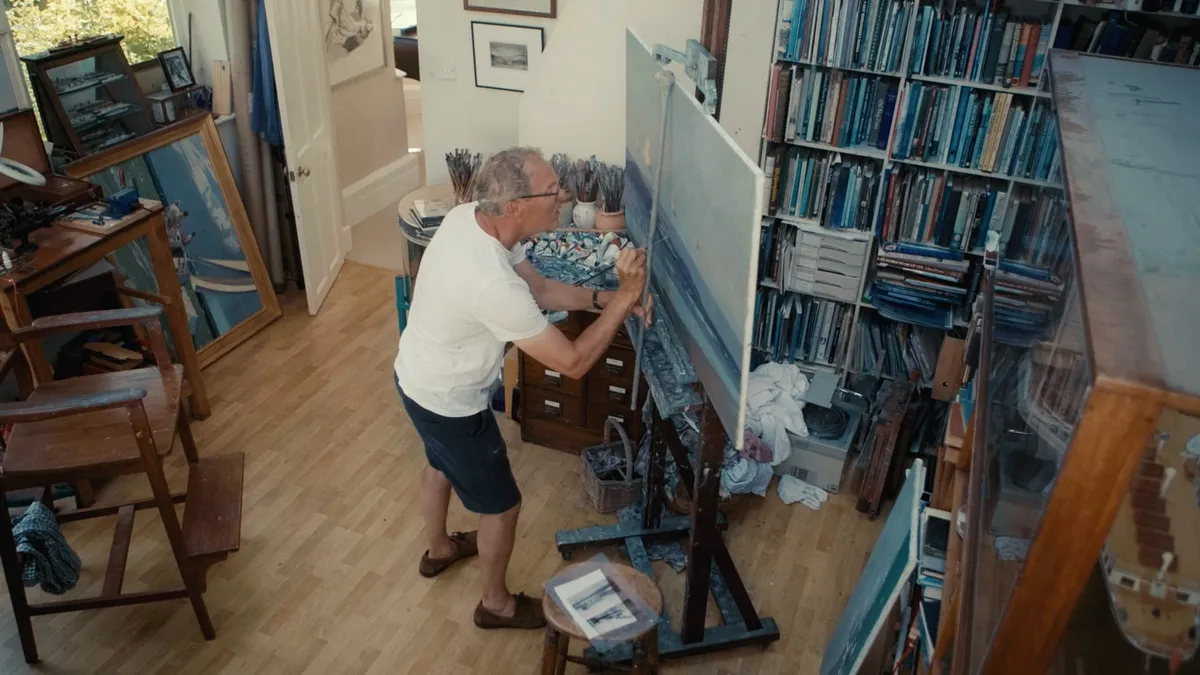 A grey-haired man in a white T-shirt and dark shorts paints at an easel, seen from above in a bookshelf-lined studio with framed pictures on the wall.