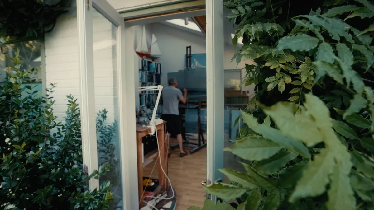 View through an open doorway framed by foliage of a grey-haired man in a white T-shirt and dark shorts painting at an easel in a wooden-floored studio.